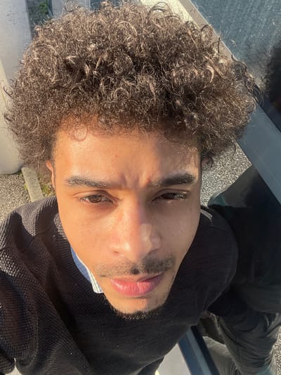 A single male individual is depicted in a high-angle, close-up photograph, likely a selfie, taken outdoors in Montigny-le-Bretonneux, France. The subject has light brown skin, dark brown curly hair, dark brown eyes, dark eyebrows, a mustache, and a goatee. He is wearing a dark, possibly black, knit sweater over a light-colored, vertically striped collared shirt. His gaze is directed towards the camera. The scene is illuminated by direct sunlight, casting highlights on the subject's face, hair, and clothing, and creating some shadowing. The immediate background to the left consists of a light grey, textured paved surface with visible aggregate and a low light-colored curb. To the right, a dark, reflective surface, consistent with a vehicle's tinted window, reflects overhead light and a portion of the subject's hair. Below this reflective surface, a dark, angled structure, possibly part of a vehicle, is visible. The ground beneath the reflective surface appears to be a darker paved material.