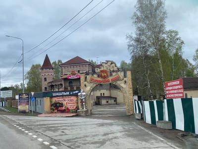 The image depicts a street scene in the city of Молодёжный, Russia, showcasing the entrance to a complex called "Рыцарский двор," which translates to "Knight's Courtyard." The entrance features a stone archway with an inscription in Russian. To the left of the entrance, there is a sign promoting a "BBQ zone" ("БАРБЕКЮ ЗОНА") offering rental services, along with contact information. There's also a smaller building with the words "ХАЛЯЛЬ ШАШЛЫК от шефа," indicating halal grilled meat from the chef.
A statue of a knight stands near the entrance. To the left of the statue, there are signs advertising construction materials (roofs, facades, etc.) and events (weddings, corporate events, birthdays). On the right side, a sign warns against blocking the entrance with snow, stating that it is private territory. A fence of green and white vertical stripes is visible next to the sign.
In the background, a building resembling a castle is visible, with a tower and brickwork. The sky is overcast, suggesting a cloudy day. The road in the foreground is wet, indicating recent or ongoing precipitation. A utility pole with wires is on the left side of the street.