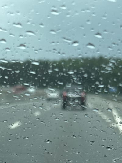 The image is taken from inside a vehicle on a wet road, looking through a rain-streaked windshield. The view is blurred due to the rain and the glass. In the background, a line of trees is visible, suggesting a semi-urban or rural setting. Several vehicles are present on the road, with one red car in the middle lane appearing most prominent. The road itself is wet and appears to be a highway or a multi-lane road. White lane markings are visible on the road. The overall lighting suggests an overcast or rainy day. The location is identified as Kerro Massiv, Russia.