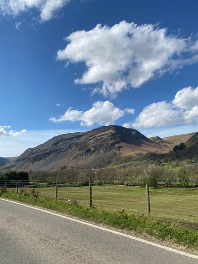 A vertical, eye-level shot depicts a landscape near Pen-y-bont-fawr, United Kingdom. A mountain with brown and gray rock formations occupies the central portion of the image, reaching from near the bottom to beyond the top center. To the left of the mountain is a bright blue sky with scattered, fluffy white clouds. To the right of the mountain is a green field enclosed by a wire fence with wooden posts. Behind the field, there are trees and possibly buildings. In the foreground, a gray asphalt road with a white line along the left edge is visible. The scene suggests a rural or mountainous area.