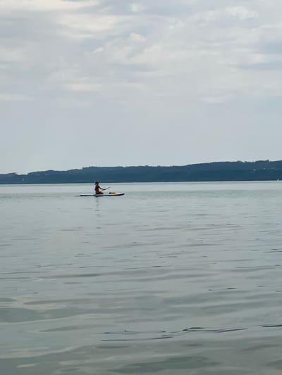 A person is paddleboarding on a lake with a tree-covered hill in the background. The sky is overcast with clouds. The water is calm and reflects the sky. The paddleboarder is wearing a bathing suit and is holding a paddle. There is a yellow bag on the paddleboard. The scene is serene and peaceful, suggesting a quiet day on the water. The location is the Lake Neuchâtel in Cortaillod, Switzerland.