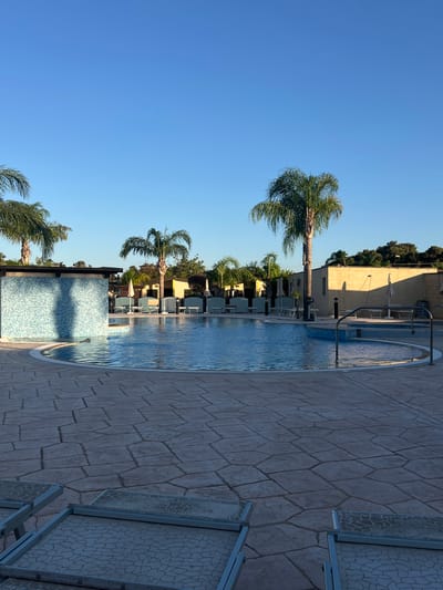 The image depicts a resort swimming pool area on a clear day in Otranto, Italy. The foreground is dominated by a large paved patio area with interlocking stone tiles of various irregular shapes and sizes. Several empty lounge chairs with patterned grey fabric and metal frames are visible on the patio, two in the lower left and two in the lower right of the frame.
The main subject of the image is a large, kidney-shaped swimming pool with clear blue water. The pool is surrounded by a white border. Part of the pool has a tiled accent, featuring small blue and white mosaic tiles, which also extends to a shallow ledge area within the pool. A metal railing with vertical supports and a horizontal bar is present along the edge of this shallow section.
Several palm trees are scattered around the pool area. Three prominent palm trees are visible in the background, with lush green fronds. Behind the pool and palm trees, there are several low-lying buildings with light-colored walls, likely part of the resort. White umbrellas are set up near some of the buildings, suggesting a seating or lounge area. The sky above is a vibrant, cloudless blue. The overall impression is a serene, well-maintained outdoor leisure space.