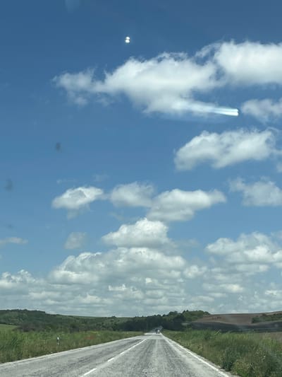 The image shows a view from a car, looking up at the sky and to the right at a road and adjacent grassy area. The sky is mostly blue with scattered white clouds. One area in the sky has a distinct, bright contrail-like formation. To the right, a straight, grey asphalt road stretches into the distance, flanked by green vegetation. A car is visible on the road in the distance. The image was taken on a roadside, at a location near Karaisen, Bulgaria.