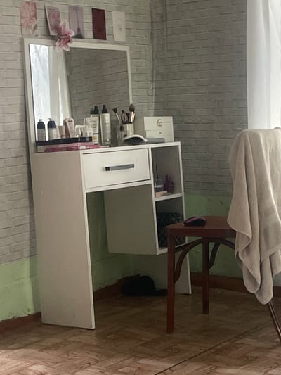The image shows a white vanity desk with a mirror. The desk is filled with various cosmetic products, including bottles of lotion, makeup brushes, and lipstick. The mirror reflects the interior of a room with a brick-patterned wall and a wooden floor. A light-colored blanket is draped over a chair next to the desk. The scene appears to be in a personal dressing area or a small salon. There are no people visible in the image. The lighting suggests it could be daytime. The text "LF" is visible on a small white object on the left side of the vanity.