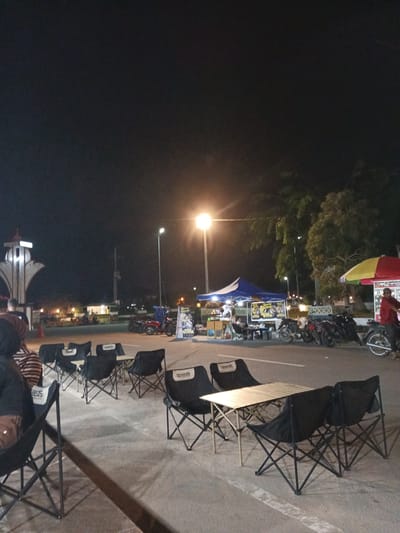 This image depicts an outdoor scene at night, likely a gathering area or food court, in Meunasahaluedrienlhoksukon Barat, Indonesia. Several folding chairs and tables are set up on a paved surface, suggesting an informal dining or seating arrangement. In the background, a large archway structure is visible, illuminated by lights.
Various food stalls are present, with one featuring a colorful umbrella and a sign that reads "Warung Keluarga." Another stall is covered by a blue tent. Several motorcycles are parked nearby, indicating modes of transportation for patrons. A few individuals are visible, either seated at the tables or walking around. One person in a red shirt stands by the "Warung Keluarga" stall. The presence of streetlights and the overall illumination suggest it is evening or night.
The scene appears to be active and social, with people gathered around the food stalls. The lighting casts a warm glow, and the atmosphere seems casual and relaxed. The ground is a concrete surface, and trees are visible in the background. The weather appears clear. The visible text includes "Speedo" on some of the chairs and "Warung Keluarga" on a food stall sign.