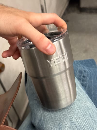 A close-up, eye-level shot features a person's hand holding a brushed stainless steel tumbler with a clear plastic lid. The word "YETI" is embossed in capital letters on the side of the tumbler. The tumbler is positioned against or near a person's leg, which is clad in light blue denim jeans. In the lower right corner, a brown leather object, possibly a shoe or part of a bag, is partially visible. The background is out of focus, showing indistinct grey and lighter tones, suggesting an indoor environment. The image is brightly lit, but the time of day is not discernible.