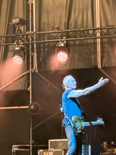 The image shows a live music performance at the Concert Music Festival in Chiclana de la Frontera, Spain. The main subject is a male musician with short, grey hair, wearing a dark blue t-shirt and blue jeans. He is actively playing a dark-colored bass guitar. His right arm is extended with his palm facing outward, as if addressing the audience.

The stage is lit with several spotlights, some of which are visible in the upper portion of the image, casting a pinkish glow. In the background, there are stage structures, lighting rigs, and fabric drapes. To the far right, a portion of another person, possibly a performer or crew member, is visible, looking towards the stage. The overall atmosphere appears to be that of an outdoor concert.