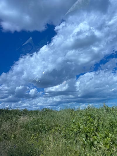The image shows a landscape with a cloudy blue sky and a field of green grass. There is a reflection on the sky that appears to be from a window, showing part of a car interior. The location is stated to be Konstantinovo, Bulgaria, but there are no specific landmarks or people visible to confirm this. The scene is natural and serene, with no discernable activity or interaction.