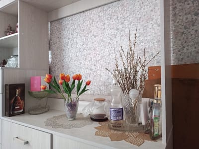 The image depicts a shelf in a domestic setting. On the shelf, there are several decorative items. To the left, a bottle of cognac is visible. In the center, a bouquet of artificial tulips is arranged in a glass vase. Behind the flowers, there's a decorative glass bowl. To the right, a plastic bottle of liquid and a tall, slender bottle with a gold cap are placed in a clear tray with a decorative pattern. A sprig of pussy willow branches is also present in the arrangement. The background features wallpaper with a floral pattern. The overall environment suggests a home interior. This scene is located in Gusinoozyorsk, Russia.