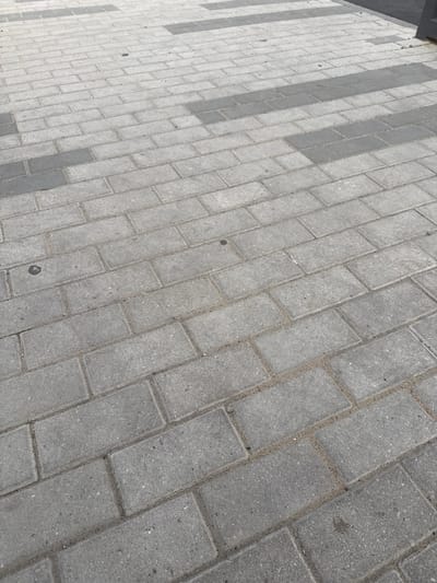 The image shows a section of a paved walkway in Koltsovo, Russia. The walkway is constructed from rectangular interlocking concrete pavers arranged in a running bond pattern. The pavers vary in color, with some being a lighter gray and others a darker gray, forming a subtle striped design across the walkway. The joints between the pavers are filled with sand. There is a dark rectangular object, possibly a bollard or a portion of a structure, visible along the right edge of the frame. The walkway appears to be outdoors, with no identifiable buildings or significant vegetation in the visible area. The lighting suggests it is daytime.