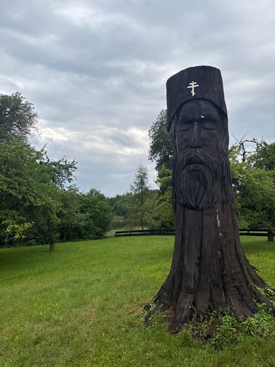 The image displays a landscape scene in Wojnowo, Poland. The primary subject is a large, dark, weathered wooden sculpture, carved from a tree trunk, positioned slightly right of center in the foreground. The sculpture depicts a male figure with a long beard, closed eyes, and a tall, cylindrical hat. An off-white Orthodox cross is affixed to the front of the hat. The base of the sculpture is integrated into a vibrant green, mown grassy field that occupies the lower half of the frame. In the midground, a dark wooden fence runs horizontally, separating the field from a body of water, likely a lake or pond, visible in the background. Dense green foliage, consisting of various trees, surrounds the water and extends towards the horizon. The sky is uniformly overcast with a layer of grey and white clouds. No human subjects are present; the scene is static.