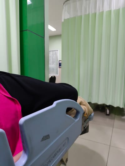 This media file depicts a close-up view of a patient lying on a hospital bed. The patient is partially visible, wearing a bright pink garment and a dark-colored covering. The bed itself is light blue with visible markings that include the text "PARAMOUNT BED". The scene appears to be within a healthcare facility, indicated by the presence of light green curtains, often used to create privacy in patient rooms or examination areas. A green pillar with a dark line accent is also visible to the left, adding a splash of color to the otherwise muted tones. The floor is tiled in a light color. In the background, through an opening, a hallway is visible with fluorescent lighting and another chair. The overall impression is that of a hospital waiting area or a patient room. No specific time of day or weather conditions can be inferred. The location is identified as Balongbendo, Indonesia. The image focuses on the immediate surroundings of the patient, suggesting a sense of stillness or waiting.