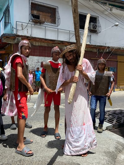 A group of people enact a religious procession on a street in San José de Barlovento, Venezuela. A man portraying Jesus Christ is carrying a wooden cross, wearing a white robe stained with red markings simulating blood and a crown of thorns. He is barefoot. Two people dressed as Roman soldiers flank him, wearing cardboard armor and helmets. A third person follows behind, also in costume. The street is lined with buildings, and a sewer grate is visible on the right side of the image.