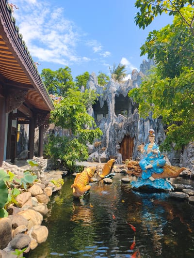 A tranquil scene unfolds in a courtyard with a pond, likely located in Tuy An Nam, Vietnam. In the foreground, a pond with dark, reflective water is bordered by large, smooth stones and lush green lily pads. Two golden koi fish sculptures are present in the pond, one on the left with water spouting from its mouth, and another to the right. In the center of the pond, a statue of a woman in a blue robe sits atop a mythical dragon-like creature. The woman has fair skin, dark hair, and is adorned with jewelry. The dragon is a reddish-brown color with its mouth open. Behind the pond, a large, artificial rock formation with arched openings and a wooden door suggests a temple or shrine. To the left of the rock formation, a traditional Vietnamese-style building with a dark wooden roof and pillars is visible. Green trees with abundant foliage surround the area, and the sky above is a bright blue with scattered white clouds.