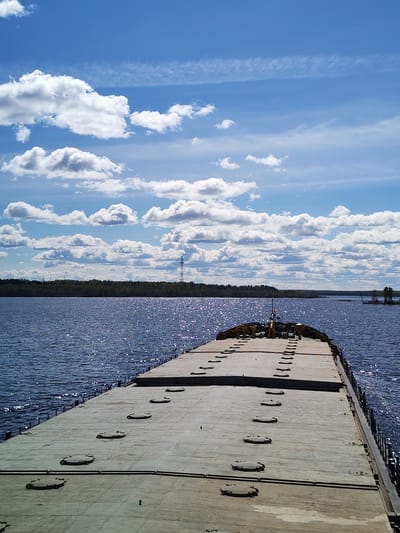 The image depicts a barge on a wide body of water, possibly a river or lake, under a partly cloudy sky. The barge is flat and grey, featuring numerous hatches. The water reflects sunlight, creating a shimmering effect. In the background, a forested shoreline is visible with an electrical transmission tower. A structure is partially visible in the far right of the image. The image is taken from a high vantage point, looking lengthwise down the barge. The location may be near Desyatovskaya, Russia, but the image alone does not confirm this location, but is taken from a waterway.