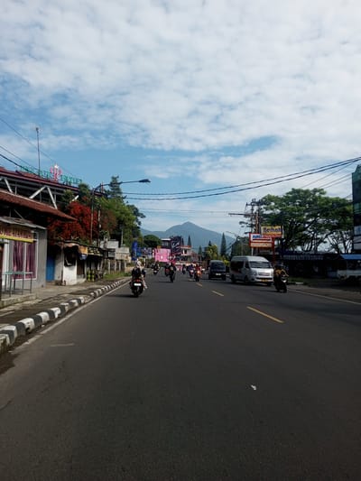 The image depicts a street scene with a mountain in the background under a partially cloudy sky. The location is likely a city or town due to the presence of buildings and commercial signage. A road with a yellow center line divides traffic. A line of motorcycles is traveling in the same direction, along with cars and a van. The roadside features buildings, including a hotel, and street-level shops. Power lines are visible overhead. The roadside edge has black and white striped curbing. The image does not appear to be in Zeyathiri District, Myanmar.
