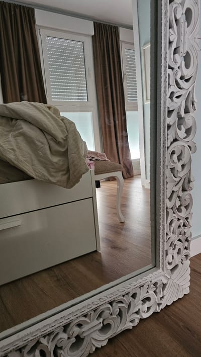 The image depicts an interior bedroom scene. A large, ornate, white-framed mirror is positioned in the foreground, reflecting a portion of the room. To the left of the mirror, a bed with rumpled, light-colored bedding is visible. Behind the bed, there are two windows with brown curtains and white roller blinds. A small, upholstered stool with white legs is situated near the bed. The floor is made of wooden planks. The room appears to be in a residential setting. There is no indication of the location being in the city of María De Huerva, Spain. The scene is static and there are no people or actions depicted.