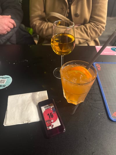 The image depicts a close-up, high-angle view of a dark, slightly reflective table surface in Saint-André-lez-Lille, France, likely within a restaurant or bar setting. In the foreground, a white, folded paper napkin rests to the left of a dark purple or black rectangular electronic device. This device displays a graphic featuring a red circle with the white number "16", the text "SMOKEY BANH MI" above it, and two cartoon characters, one red and one blue. To the right of these items, a faceted glass tumbler contains an orange-colored beverage with ice cubes and an orange slice garnish, penetrated by a dark-colored straw. Further to the right and slightly behind, a clear stemmed wine glass holds a light amber liquid. In the blurry background, two individuals are partially visible: one on the left with a dark sleeve and hand, and another in the center-right wearing a light brown buttoned coat. A light blue circular coaster with a QR code is partially visible on the far left, and a blue rectangular placemat with a grid pattern and red text is on the far right. The table shows minor smudges or spills. No specific actions or interactions are discernible.