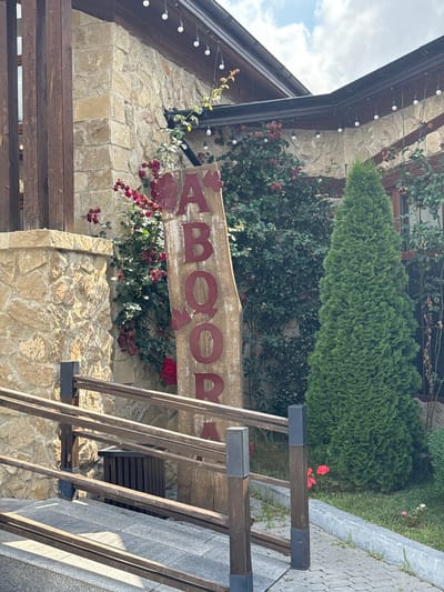 A weathered wooden sign with red lettering stands vertically near the entrance of a building. The letters spell out "AABOOR," with the last two letters partially obscured by foliage. The building's exterior is constructed of rough, light-colored stone blocks, and dark wooden beams are visible along the roofline and supporting the structure. Strings of small, round lights are strung along the overhang of the roof. Lush green foliage, including climbing plants and a tall, conical evergreen tree, surrounds the building. Red flowers bloom amongst the greenery. A wooden railing with horizontal planks leads up a ramp to the entrance. The ground is paved with grey stones and concrete. The scene appears to be an outdoor area of a establishment in Mirikənd, Azerbaijan.