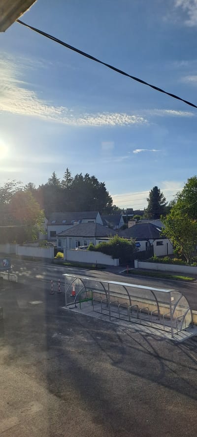 The image captures a street scene in Castlebar, Ireland, bathed in the soft light of a sunny day. A modern, transparent-roofed bicycle shelter with metal racks stands prominently in the foreground, casting long shadows on the asphalt. Adjacent to it, traffic cones and barriers indicate ongoing work or a restricted area. Across the street, a low, white-walled barrier encloses several houses with pitched roofs, typical of suburban Irish architecture, some with dormer windows. Lush green trees and foliage surround the properties, with taller conifer trees forming a dense backdrop against a bright blue sky dotted with wispy white clouds. The sun, low in the sky, creates a slight glare on the left side of the image. No people are visible in the frame.