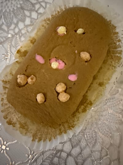 The image shows a close-up of a brown, oval-shaped pastry with light yellow and pink frosting dots and small, light brown pieces resembling cereal or nuts. The pastry is sitting in a white, fluted paper liner, which is partially resting on a white, patterned fabric. The background is a blurry, light-colored surface with an ornate, possibly lace-like, pattern. There is no discernible urban environment or specific location information present in the image.