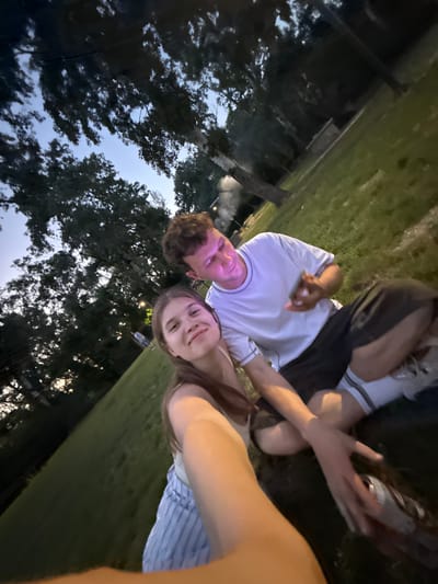 A photograph taken in Cukarica Urban Municipality, Serbia, at twilight shows a young man and woman reclining in a grassy area near trees. The man is wearing a white shirt and the woman is wearing a striped top. The photo is taken from a low angle and rotated counterclockwise.