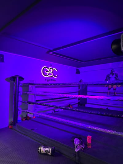 This media file depicts a boxing ring inside a dimly lit, neon-lit gym. The scene is bathed in purple and pink hues from the ambient lighting, creating an intense atmosphere. The camera is angled from the top of the ring looking down, with the ropes of the ring prominently featured in the foreground.

Visible objects include boxing gloves, a water bottle, and possibly some training equipment. On the wall behind the ring, there is a large stylized logo that reads "GBC Fight Club". A graphic of a boxer is also visible on the wall. The overall setting suggests a dedicated space for boxing training or competition.

There are no people visibly present in the frame, but the presence of the equipment and the branded signage implies that the location is actively used for its intended purpose. The time of day is indeterminate due to the artificial lighting, and no weather cues are present. The scene evokes a sense of determination and grit associated with the sport of boxing. The location context is Kâğıthane, Turkey.