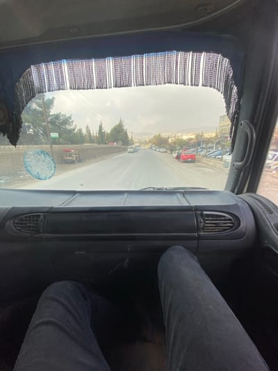 The image depicts a first-person perspective from inside a vehicle looking forward through the windshield onto a road in Dahiyat Harasta, Syria. The foreground shows the top of a dark dashboard featuring two air vents, and two legs clad in dark blue jeans extended into the lower part of the frame. A dark, fringed curtain hangs along the top edge of the windshield.
The exterior scene reveals a two-lane asphalt road extending into the distance under an overcast sky. The road surface appears damp. On the left side, a concrete barrier runs parallel to the road, beyond which mature green coniferous trees are visible, along with a utility pole supporting power lines. A small, light-colored vehicle with a red canopy is parked behind the barrier. A faded, circular blue adhesive or mark is present on the lower-left portion of the windshield. On the right side of the road, multiple vehicles are parked along a shoulder or unpaved area, including a prominent red pickup truck. Further down, buildings suggestive of commercial or residential structures line the road. In the far distance, hills or mountains are visible beneath the cloudy sky. Sparse traffic is present on the road ahead.