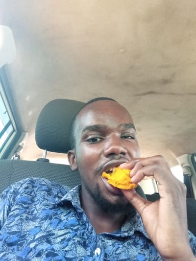 A single adult male is depicted from the chest up, situated inside a vehicle in the Ganjoni/Shimanzi ward, Kenya. The individual has dark skin, short dark hair, and a short beard. He is wearing a blue short-sleeved collared shirt featuring a light-colored abstract pattern. The man is actively consuming a bright orange or yellow, irregularly shaped food item, which he holds to his mouth with his right hand. His mouth is open, revealing his teeth as he bites into the item. Perspiration is visible on his forehead and upper lip. He is looking directly into the camera. The vehicle's interior includes a light-colored, possibly stained, fabric headliner and a dark grey textured fabric seat with a headrest positioned behind the man. A light-framed window is visible on the left side of the frame.