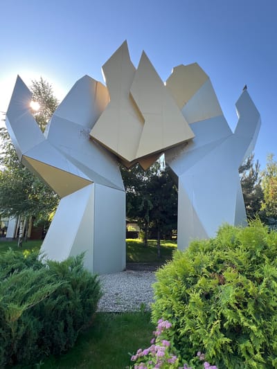 A large, abstract sculpture made of metallic, angular panels stands outdoors. The sculpture is primarily silver and gold, with some darker areas that may indicate weathering or shadow. It has a complex, geometric design that resembles large, interlocking hands or abstract wings reaching upwards. The sculpture is situated in a park-like setting with green grass, lush bushes in the foreground, and trees in the background. The sun is shining brightly, casting lens flare through the sculpture and illuminating the scene. A bird is perched on the upper portion of the sculpture. The ground beneath the sculpture appears to be covered in gravel. The overall impression is of a modern art installation in a natural environment. This sculpture is located in Plastunivska, Russia.