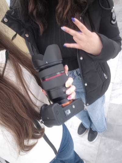 A first-person perspective photograph shows two people indoors on a white tiled floor. The person taking the photo is wearing blue jeans and black slip-on shoes, and holding a black digital camera with a large lens in their left hand. Their nails are painted with various colors. They are also wearing a black puffer jacket over a black shirt. The other person is wearing blue jeans and a white top. Both people have brown hair. The location is likely indoors, as the floor is tiled. Given the request for a location in Srebarna, Bulgaria, and the absence of external environmental cues, this photo could be taken in a house, hotel, or other indoor facility in Srebarna.