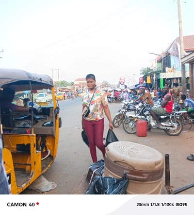 An eye-level, medium shot reveals a bustling street scene. A woman in a patterned shirt and maroon pants walks towards the camera, carrying black bags. Parked beside her is a yellow auto rickshaw. Motorbikes, each with a rider, line the street further ahead. A large advertisement featuring men in suits is visible in the background, above the crowd of people. The street scene is lively and filled with activity. A large, brown trash can sits next to the woman, and the lighting is bright and even.