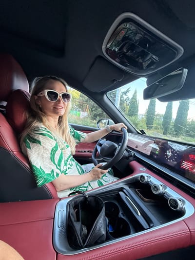 A blonde woman with blonde hair and sunglasses is driving a car. She is wearing a green and white floral print dress. The car interior is red and black. The car has a large digital dashboard. Outside the car, there are trees and a blue sky. The image is taken from inside the car, looking towards the driver. The location is Pushkino, Ukraine.