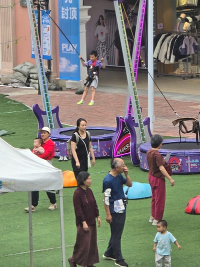 In Fuyang, China, an outdoor scene shows a young boy in a purple harness attached to a bungee trampoline. He is mid-air, his legs extended. The trampoline area has a large purple guitar-shaped prop with neon lights. Several other people are present on a green artificial turf. A woman in a red shirt holds a baby, standing near a white tent. Another woman in a black t-shirt and white backpack walks by. An older couple, a man in a blue shirt and a woman in a brown top, are also in the frame. A young child in a light blue t-shirt stands in the foreground. In the background, shops with clothing racks are visible.