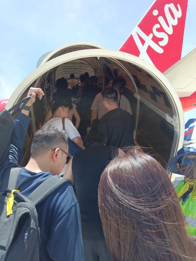 An outdoor, eye-level medium shot captures a group of airline passengers boarding an airplane via a mobile staircase under a clear blue sky. The red tail fin of an AirAsia plane is visible in the upper right corner. The staircase is a curved, metallic structure with a dark interior. People are visible ascending the stairs, some carrying bags. In the foreground, the back of a man's head wearing sunglasses and a navy blue shirt is prominent, with a black backpack slung over his shoulder. To his right, the back of a woman's head with long, dark brown hair is visible. Further back, several other individuals, mostly men, are seen ascending the stairs, dressed in casual attire. The overall scene suggests the process of boarding an aircraft. This scene is taking place at Cam Ranh International Airport in Cam Ranh City, Vietnam.