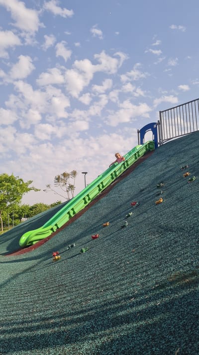 A photograph taken from below and angled upward depicts a climbing wall structure against a blue sky with scattered clouds. The climbing wall is surfaced with a green, textured material dotted with various colored climbing holds. A green slide is attached to the wall. A young girl is positioned midway down the slide, facing downward. The climbing wall is located in Kiryat Gat, Israel. Trees and greenery are visible at the base of the climbing wall. Metal safety bars are positioned at the top of the structure.