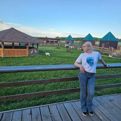 A woman with blonde hair and glasses is standing on a wooden deck, leaning on a railing. She is wearing a white t-shirt with a blue graphic and blue jeans. In the background, there is a grassy field with several small wooden buildings with teal roofs. A white goat is visible in the field. The sky is clear and blue, suggesting it is daytime with fair weather. The scene appears to be a rural or farm setting. Visible text on a building in the background reads "Бабушкин сеновал" (Babushkin senoval), which translates to "Grandmother's hayloft."