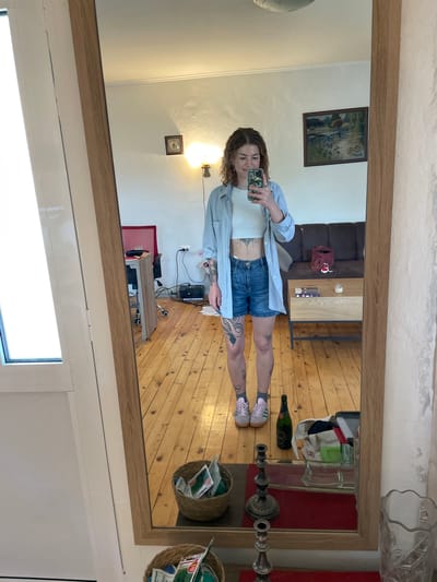 A young woman with curly brown hair is captured in a full-body mirror selfie, taken from inside a room in Danilovgrad, Montenegro. She is wearing a white crop top, denim shorts, and a light blue unbuttoned long-sleeved shirt. Her feet are clad in light pink sneakers with white laces and gray socks. Visible tattoos adorn her midriff, arms, and legs. She holds a smartphone with a patterned case in her right hand, angled to take the photo, and has a neutral to slightly smiling expression.

The mirror reflects a casual indoor setting, likely a living area, with light wooden floorboards and white walls. In the background, a dark brown sofa is visible, along with a light wood coffee table featuring metal legs. On the wall above the sofa hangs a framed landscape painting. To the right of the woman, a tall, ornate metal candelabra or lamp base stands on a reddish surface, possibly a rug or table. A dark green bottle lies on the wooden floor, near a white shopping bag. Further back, two woven baskets containing papers or magazines are visible, possibly mounted on the wall or placed on furniture. Another light wood and metal table with a black electronic device is also reflected. A wall sconce illuminates a section of a white wall, and a smaller framed picture or clock is visible nearby. The lighting suggests it is daytime. No clearly legible text is visible, though some text appears on the papers within the baskets and possibly on the sneaker tongues.