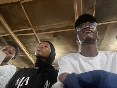 Three individuals are looking up, positioned beneath a rustic, unfinished ceiling with exposed beams. A young woman on the left has short hair and wears a light, patterned top. The woman in the center wears a dark hijab and a black shirt with white text. The man on the right wears a white t-shirt, a black baseball cap, and aviator-style glasses. He looks upward, with his mouth slightly open. The lower part of his body is covered with blue clothing.