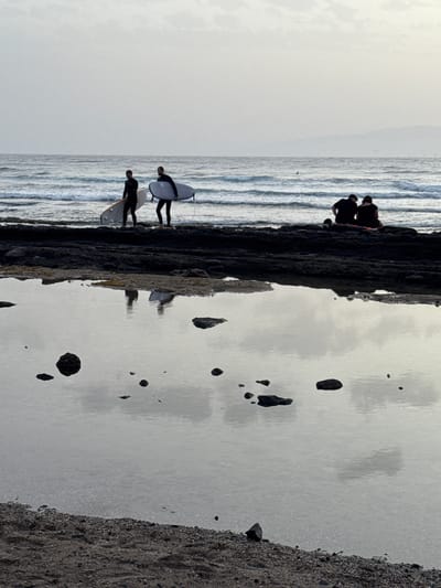 The image shows a rocky coastline with several people. In the foreground, a shallow tide pool reflects the sky and the surrounding rocks. Two surfers in wetsuits are walking on the dark rocks near the water's edge, each carrying a surfboard. Further back, two other individuals are seated on the rocks, facing the ocean. The ocean itself is calm with gentle waves. In the distance, a faint outline of land is visible on the horizon. The overall lighting suggests it is early morning or late evening. The scene is consistent with a location along the coast of Playa De Las Américas, Spain, known for its volcanic rock formations and surf spots.