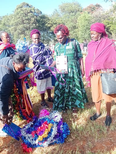 Five women stand outdoors looking at items on the ground, possibly gifts. One woman wears a green patterned dress and a lanyard. Another is draped in a purple blanket over shorts. An older woman wears a maroon headscarf and shawl with a purse over her shoulder. The other women are wearing similar garments. Tinsel, wrapped gifts, and colorful decorations are scattered on the ground. Trees are visible in the background.