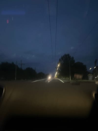The image captures a view from inside a vehicle traveling at night on a road in Strumivka, Ukraine. The sky is a deep twilight blue, with dark clouds indicating a potentially overcast or stormy evening. Overhead, power lines are visible against the sky. The road ahead is illuminated by the headlights of the vehicle and the streetlights lining the right side of the road. Trees and some structures are dimly visible on either side of the road, partially obscured by darkness and motion blur. The foreground of the image is dominated by the interior of the vehicle, with the dashboard and windshield providing a dark, blurry frame. One set of headlights from an oncoming vehicle is visible in the distance.