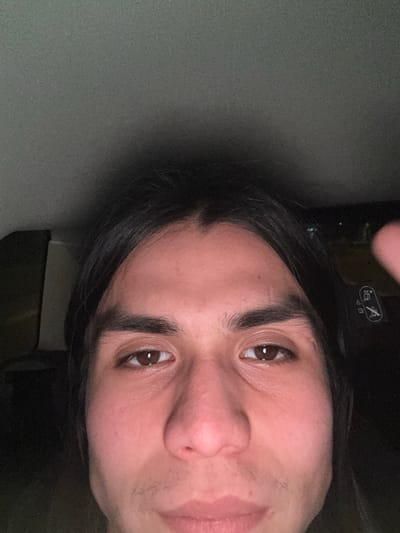 The scene, located in Riorges, France, depicts a close-up, frontal view of a single individual, male, from the forehead to the lower lip. The subject has dark, center-parted, shoulder-length hair, dark brown eyes, and a neutral facial expression. Subtle stubble is visible on the upper lip and chin. The individual is situated within what appears to be a vehicle interior; a light grey headliner occupies the top of the frame. The immediate background behind the subject is dark, with a seat or headrest component displaying white symbols visible to the upper right of the subject's head. Low-level illumination highlights the subject's face, while peripheral details within the vehicle are indistinct due to dim lighting.