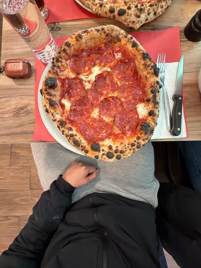 An overhead view captures a dining table within an indoor restaurant setting located in Corminboeuf, Switzerland. The central subject is a round, Neapolitan-style pizza, approximately 12 inches in diameter, served on a white plate placed over a red paper placemat. The pizza exhibits a visibly charred crust with leopard-spotting, topped with melted white cheese and numerous slices of red pepperoni or salami.

To the right of the pizza, a silver-colored fork and a silver-bladed knife with a black handle are laid on the placemat. In the upper-left quadrant of the frame, a clear glass filled with water is positioned next to a partially visible second glass containing a dark beverage. A brown, textured case, consistent with wireless earbuds, rests on the wooden table surface near the water glass.

The lower portion of the image shows the upper torso and lap of an individual, dressed in a grey sweatshirt and a black zippered jacket, with their left hand resting on their lap. A second person's leg, clad in dark blue denim fabric, is partially visible in the lower right, suggesting another diner seated at the table. The table surface is made of light-colored wood planks, and the visible floor beneath the table is also wooden.