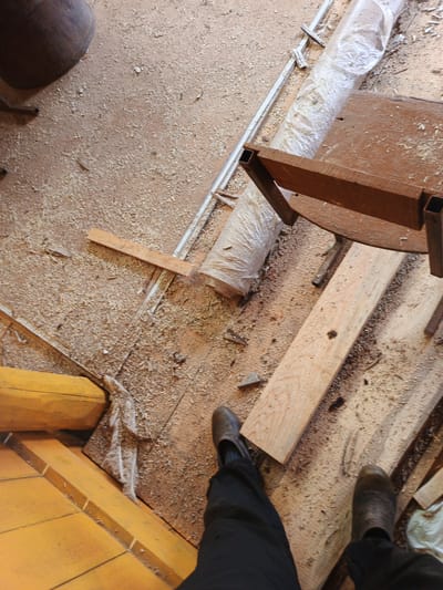 This image captures a floor covered in sawdust, likely in a workshop or construction area, located in the city of Islambaevo, Russia. A dark-colored, large cylindrical object, possibly a drum or container, is visible in the upper left corner. To the right of this object, a long, white, plastic-wrapped item is coiled on the floor. A rustic wooden table with metal legs is positioned in the mid-ground, with several wooden planks lying on and around it. In the foreground, a yellow-painted wooden structure, possibly part of a floor or wall, is visible on the left. The lower portion of the image shows a person's legs and feet clad in dark pants and boots, standing on the sawdust-covered floor. The overall environment appears to be utilitarian and reflects an active work area.