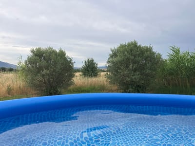 The image depicts a close-up view of the edge of a blue inflatable swimming pool filled with clear water. The water has a mosaic pattern at the bottom and ripples on the surface. In the background, there are green trees, dry grass, and tall reeds. In the far distance, mountains are visible under a cloudy sky. The scene appears to be in an outdoor, possibly rural, setting. No people or specific landmarks of Γαλλικός, Greece are identifiable in the image.