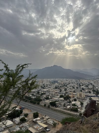 The image is a high-angle, vertical-oriented shot of Khor Fakkan, United Arab Emirates, taken on an overcast day with sun rays piercing through the clouds. Mountains form a backdrop to the cityscape. A road runs along the edge of the city, with buildings densely packed together. Vegetation is visible in the foreground and interspersed within the urban area. The shot seems to be taken from a vantage point on a slope or hill overlooking the city.