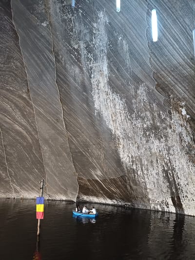 The image depicts an underground salt mine in Turda, Romania. A dark, still body of water fills the foreground, with a small blue boat carrying three people gently navigating the surface. The boat's occupants appear to be engaged in conversation or enjoying the experience. To the left, a wooden pole stands in the water, adorned with the Romanian flag, its colors of blue, yellow, and red clearly visible.
The background is dominated by the cavernous walls of the salt mine, characterized by dramatic, stripy rock formations and patches of white salt deposits. The sheer scale of the mine is emphasized by the low vantage point from the boat, making the walls appear immense and imposing. The lighting, provided by several long, cylindrical fixtures hanging from the ceiling, casts a cool, white glow, illuminating the textured surfaces of the salt and rock. The overall atmosphere is serene and awe-inspiring, highlighting the unique natural beauty and industrial history of the location. The scene appears to be set during daylight, though the underground environment creates a subdued illumination.