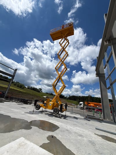 The image depicts a construction site scene under a partly cloudy sky. Dominating the center is a tall yellow scissor lift, extended upwards, with two individuals visible in the platform at the top. To the right, a section of a grey steel building frame is under construction. In the background, a hilly landscape covered with trees is visible, along with a white truck and an orange crane or similar heavy machinery. The foreground shows a wet concrete surface with puddles reflecting the surroundings. The overall impression is one of ongoing construction work in an outdoor, somewhat rural setting. The location is Janowice, Poland.