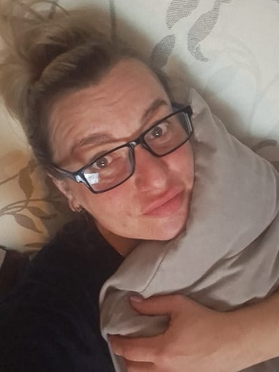 An adult female is depicted in a close-up, selfie-style photograph, lying down indoors. Her light brown hair is gathered in a loose bun on top of her head. She wears black-framed rectangular glasses, through which her wide-open eyes make direct contact with the viewer. Her lips are slightly pursed. She exhibits horizontal lines on her forehead. A dark-colored garment is visible on her upper torso, along with a thin gold chain around her neck and a small gold hoop earring on her left ear. Her right hand, displaying light pink nail polish, clutches a light grey pillow against her upper body. The background consists of light-colored bedding with a recurring pattern of dark grey or beige leaves. This scene is located in the city of Yegoryevsk, Russia.